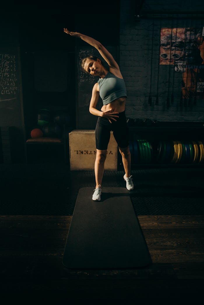 A woman performing a stretching exercise on a yoga mat in a dimly lit gym setting.